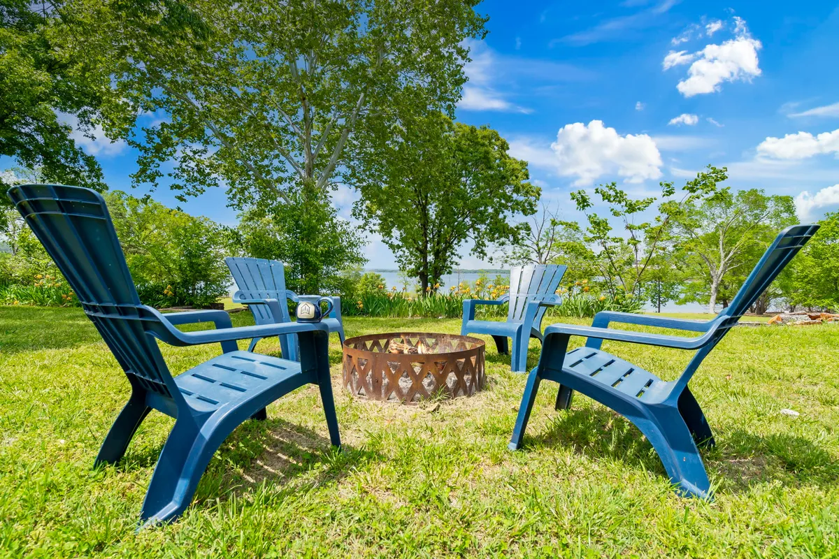 This image shows a cozy outdoor fire pit area surrounded by blue Adirondack chairs, set against lush greenery and a clear sky. The scene evokes a sense of togetherness, relaxation, and the enjoyment of outdoor evenings.