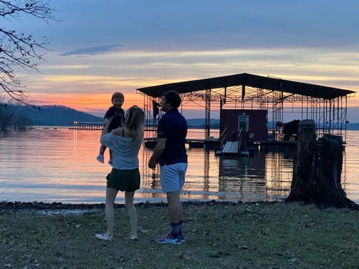 A joyful family plays along the shoreline beside the lake, with a boathouse in the background and the warm glow of sunset reflecting on the water. The scene is lively and full of energy, capturing the essence of making memories by the lake.