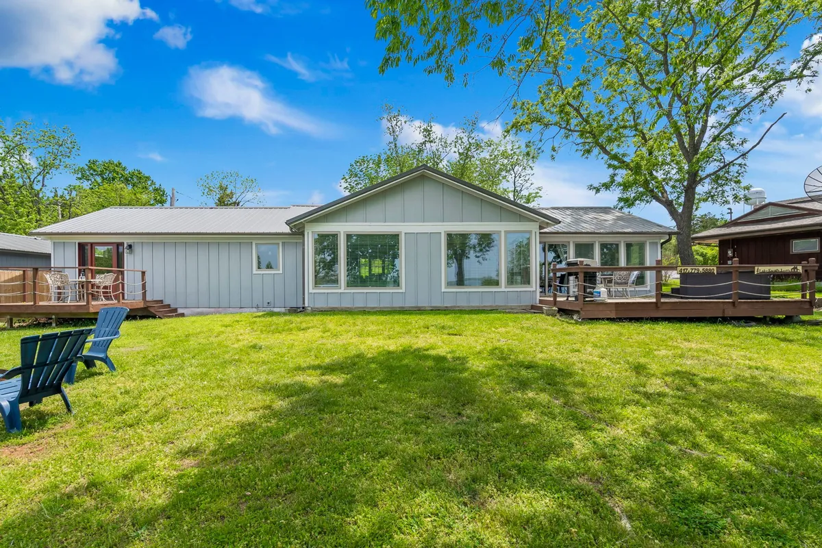 A spacious backyard with a lush green lawn framed by mature trees, featuring a large deck area with comfortable outdoor seating. The home's expansive windows overlook the yard, inviting a sense of seamless indoor-outdoor living. The setup is ideal for pets to play and families to relax together in the open air.