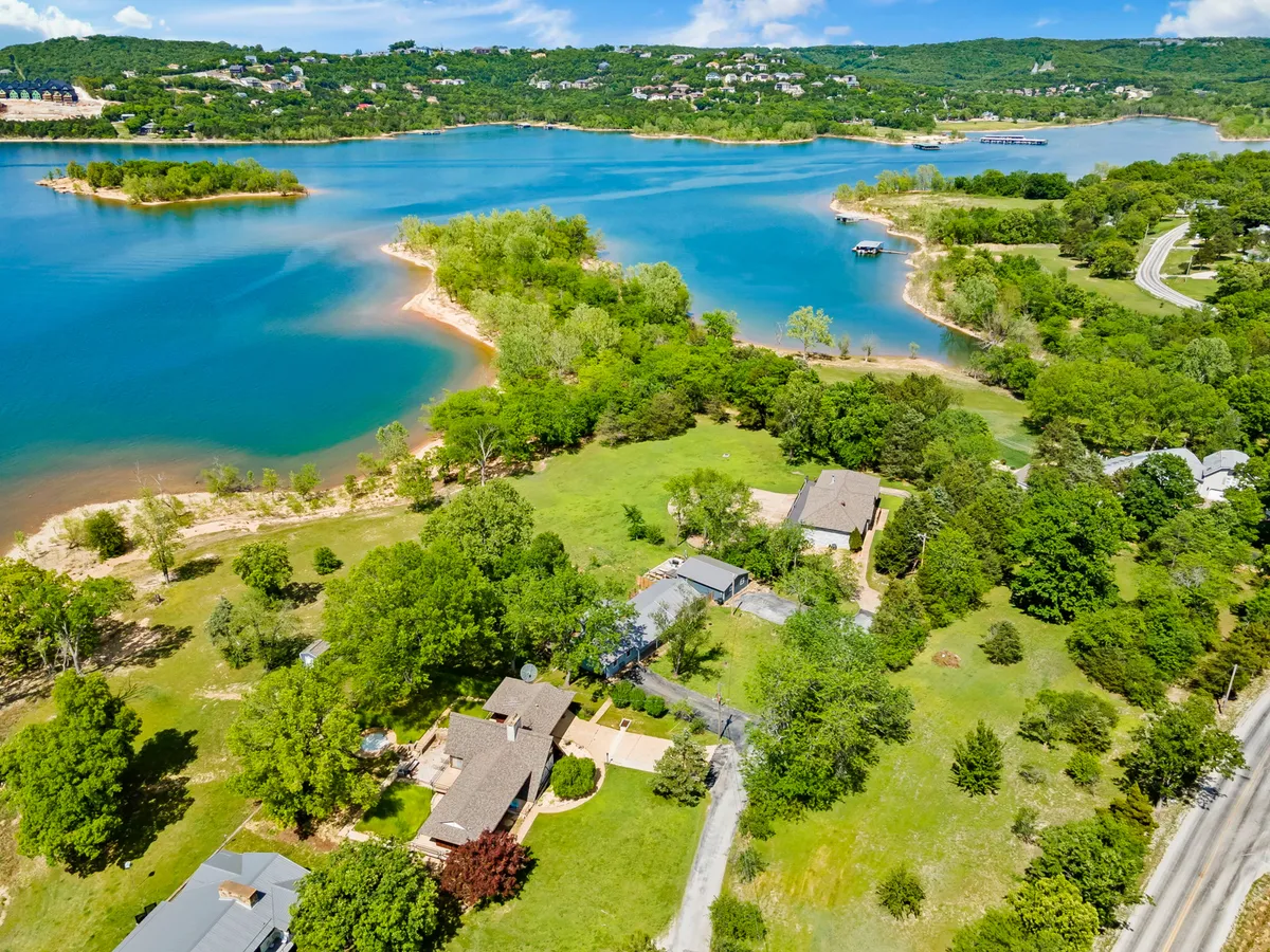 A broad aerial view showcasing the lake, nearby homes, surrounding greenery, and a road leading into the area. The composition highlights how the property nestles within nature, yet connects easily to outside attractions and roads.