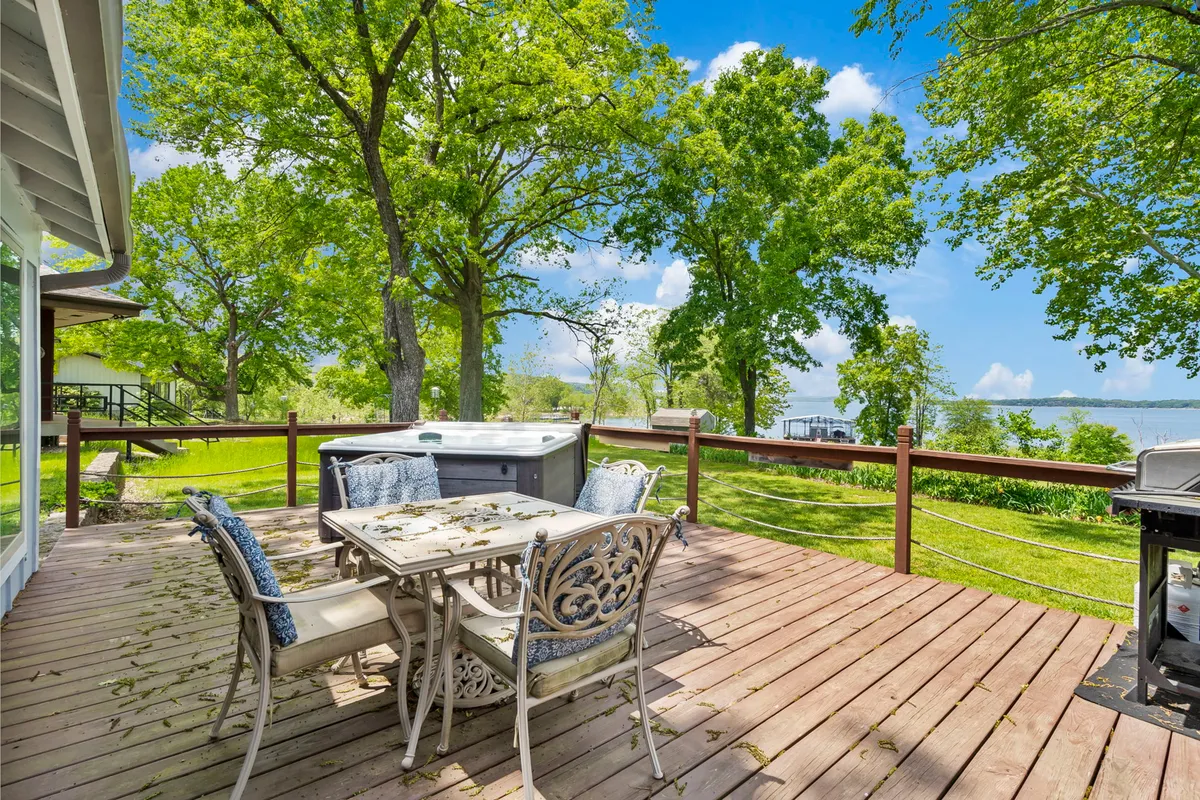 A lakeside deck features a dining table and a relaxing hot tub, overlooking the shimmering lake and surrounded by green trees. The setup is ideal for outdoor dining, morning coffee, or unwinding in the hot tub while immersed in natural beauty.