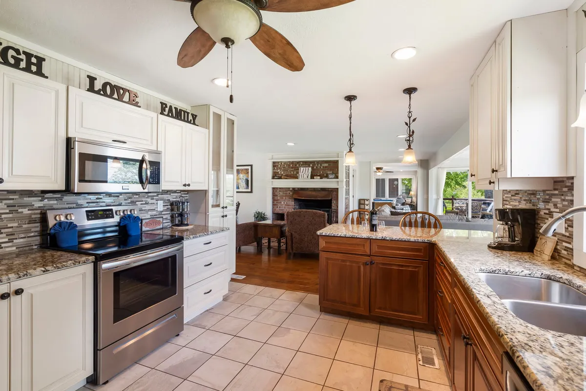 A spacious, open-concept kitchen featuring a large island with granite countertops, stainless steel appliances, and warm wooden cabinetry. In the background, a cozy living area with a fireplace creates an inviting atmosphere, ideal for gatherings and entertaining. The kitchen layout encourages interaction and is perfect for preparing meals with family or friends present.
