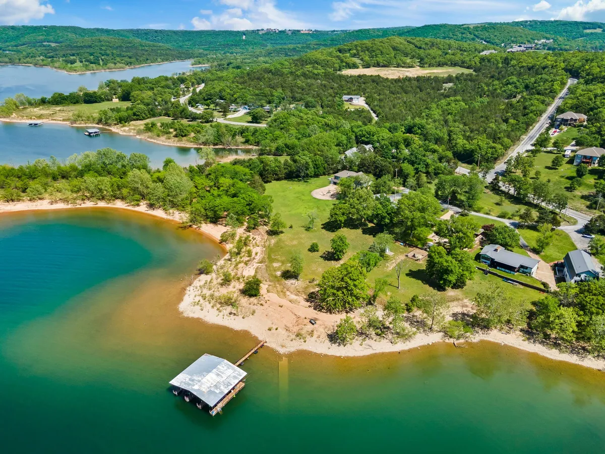 A serene lakeside setting with a dock extending over calm waters, surrounded by greenery and homes. Visible trails and open space along the shore provide ample opportunity for scenic walks and lakeside adventures with a pet.