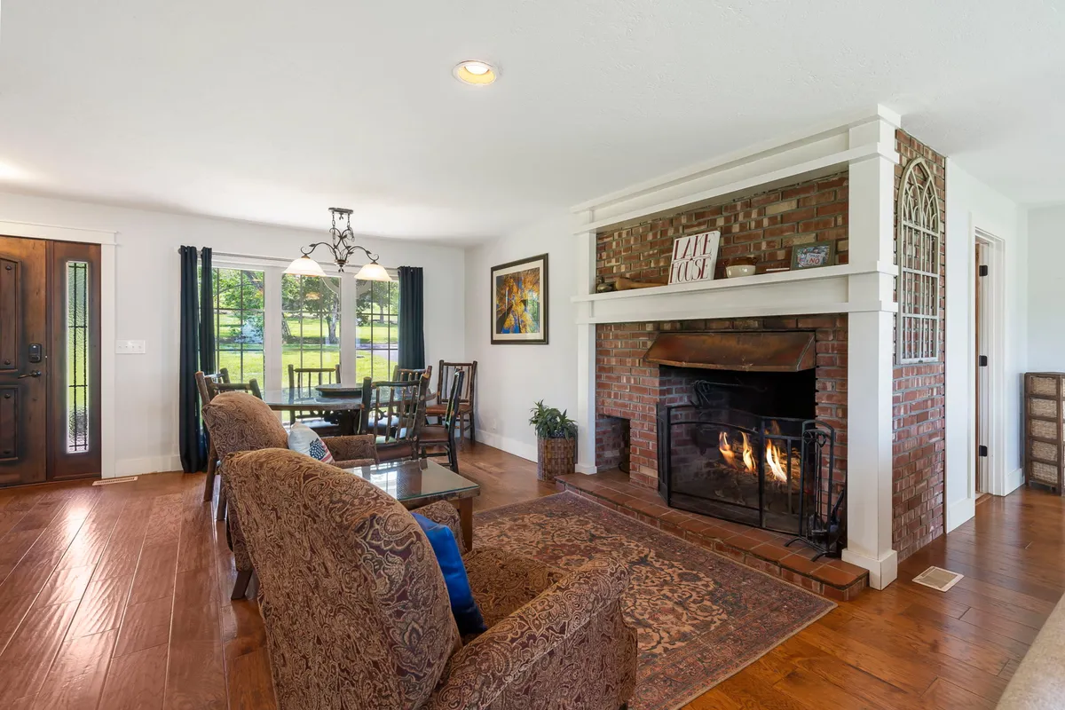 The image showcases a warm, inviting living and dining space with a glowing fireplace as the centerpiece. Large windows frame serene lake views, flooding the room with natural light, while comfortable sofas and a spacious dining table invite family gatherings and relaxing evenings. Tasteful artwork and refined finishes complete the cozy, upscale ambiance.