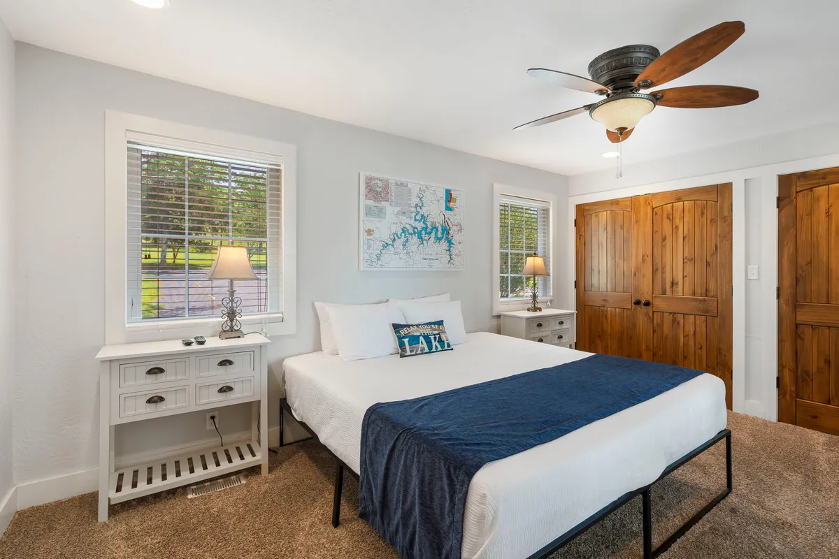 A cozy king bedroom features a bed with a plush blue throw blanket, flanked by wooden nightstands and framed by a large window. Map artwork enhances the room’s welcoming vibe, while the window ensures plenty of natural light for a bright, inviting space.