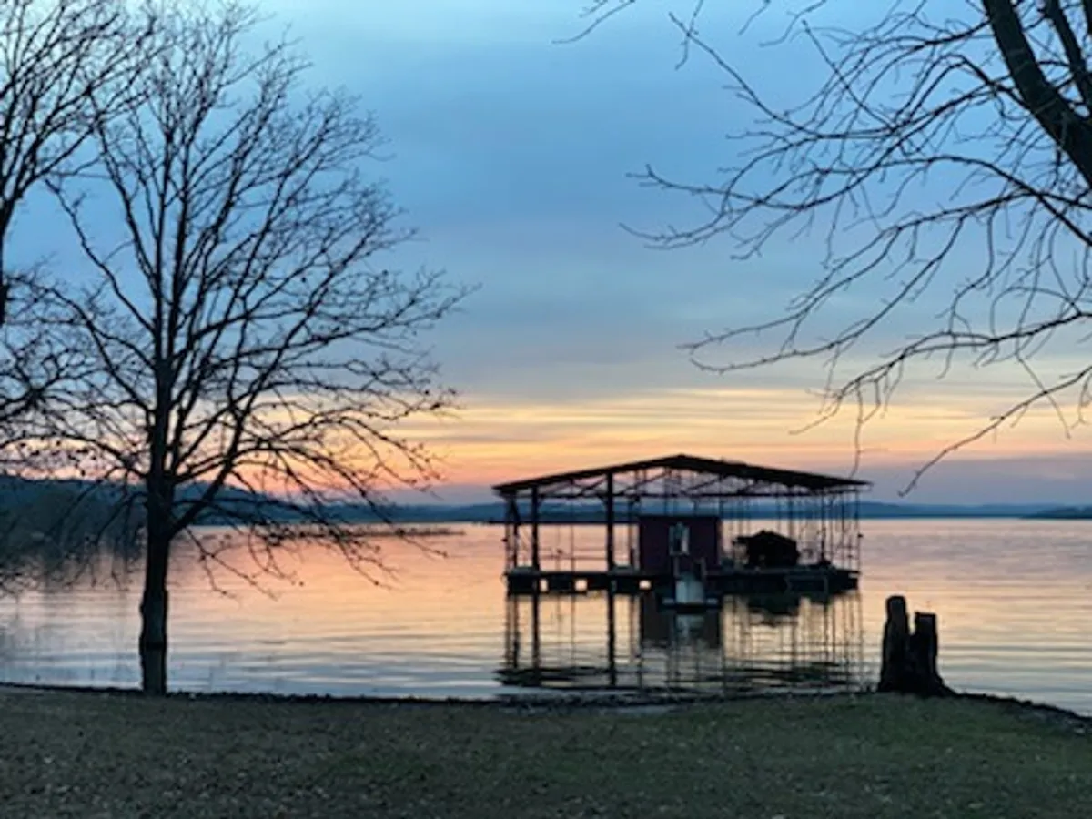 A serene dock stretches out over the calm waters of Table Rock Lake, with a vibrant sunset sky reflected in the glassy surface. Trees frame the scene, and the dock invites activities like fishing, launching kayaks, or simply relaxing close to the water.