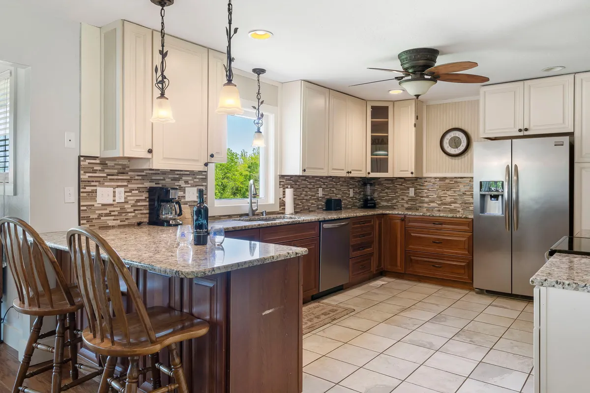 A close-up view of the kitchen’s granite countertop, accompanied by wooden bar stools and a stylish mosaic backsplash. Stainless steel appliances and a window with views of the outdoors are also visible, setting a scene for casual dining and socializing at the bar top.