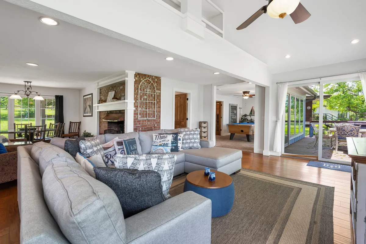 This image captures the heart of the lodge’s open plan living area: a spacious sectional sofa faces a warm wood-burning fireplace, and large windows look out onto the lake. The dining area and a pool table are also visible, reinforcing the sense of communal gathering and fun. Sunlight floods the space, emphasizing the connection to the outdoors.