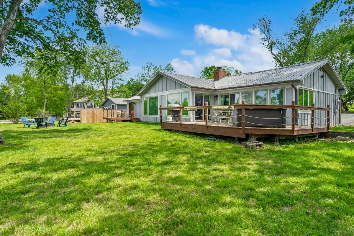 A cheerful exterior scene with a deck area furnished with seating, a large green lawn, and surrounding trees. The space looks ideal for outdoor relaxation, family barbecues, and nature walks. The open access between the home and yard is perfect for pets to stretch their legs and for guests to enjoy lakeside breezes.