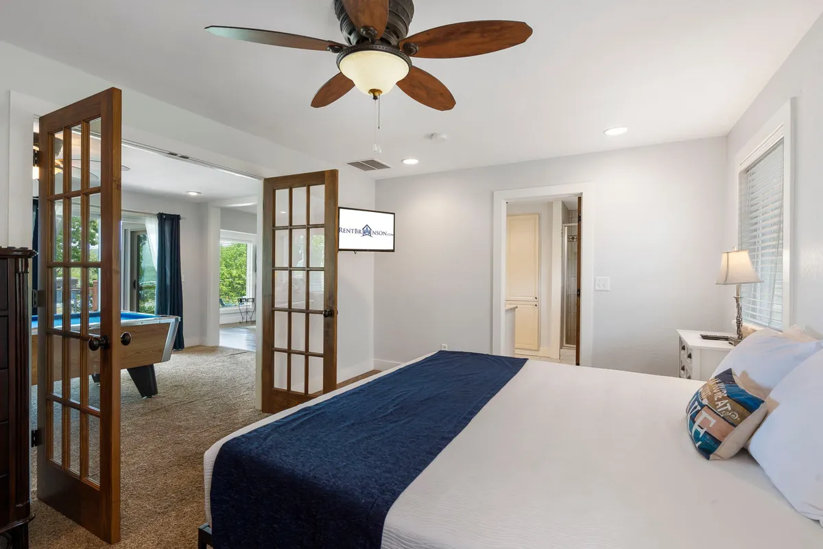 A spacious king-sized bed is the focal point of this tranquil bedroom, accented by French doors that let in ample natural light. A ceiling fan hangs above for comfort, and a decorative pillow adds a touch of style. The serene setting is ideal for restful sleep and quiet mornings, with the promise of lake views just beyond the doors.