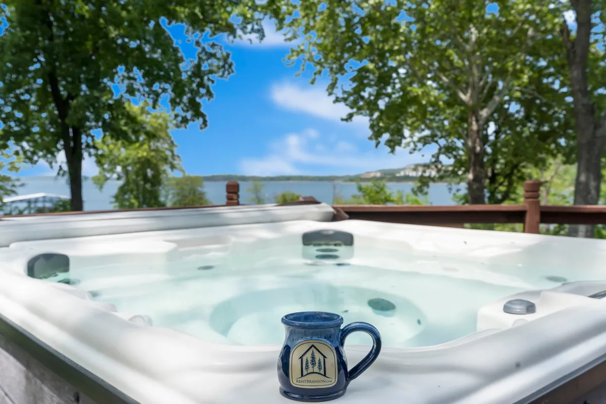 A steaming hot tub sits on a deck with a mug resting on its ledge, surrounded by trees and overlooking the lake. The tranquil atmosphere suggests both relaxation and the ability to enjoy the outdoors in cooler months.